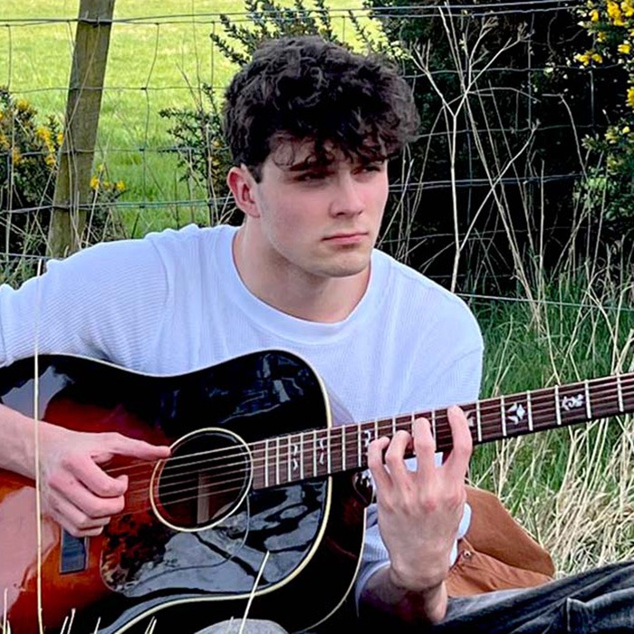 TomSmallHP2 Tom Rhodes sitting in a field with a brown acoustic guitar